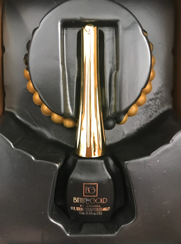 the inside of the Bitter Gold Nail Serum box with the black and gold bottle and silicone anti-stress bracelet, neversaydiebeauty.com