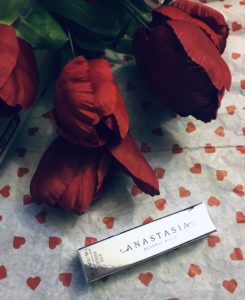 shiny silver foil box of Anastasia Beverly Hills Eye Primer against heart paper background with red tulips